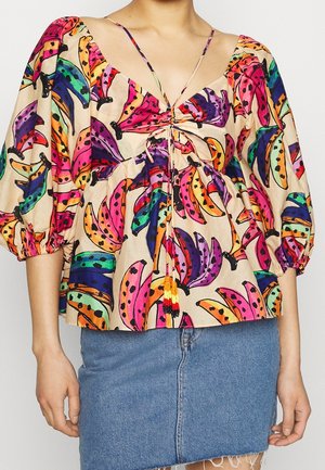 Femme portant un chemisier coloré à manches bouffantes avec un imprimé bananes et un détail de nouage perlée, associé à une jupe en jean bleue.