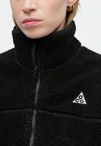 Jachetă din polar neagră cu guler înalt și închidere cu fermoar. Prezintă un patch cu logo și un material texturat. Detalii cu fermoar sunt vizibile pe față.