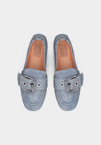 Ζευγάρι ανοιχτόχρωμων μπλε τζιν μοκασίνια με μεγάλες ασημένιες αγκράφες και ραμμένες λεπτομέρειες, απεικονισμένα από πάνω πάνω σε λευκό φόντο.