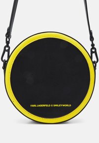 Kulatá crossbody taška s černým vnějším pláštěm, žlutým lemem a kontrastním šitím. Obsahuje detaily loga a řetízkový popruh.