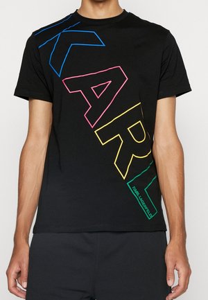 Hombre con una camiseta negra que tiene grandes letras contorneadas "KARL" en azul, rosa, amarillo y verde en diagonal en el frente.