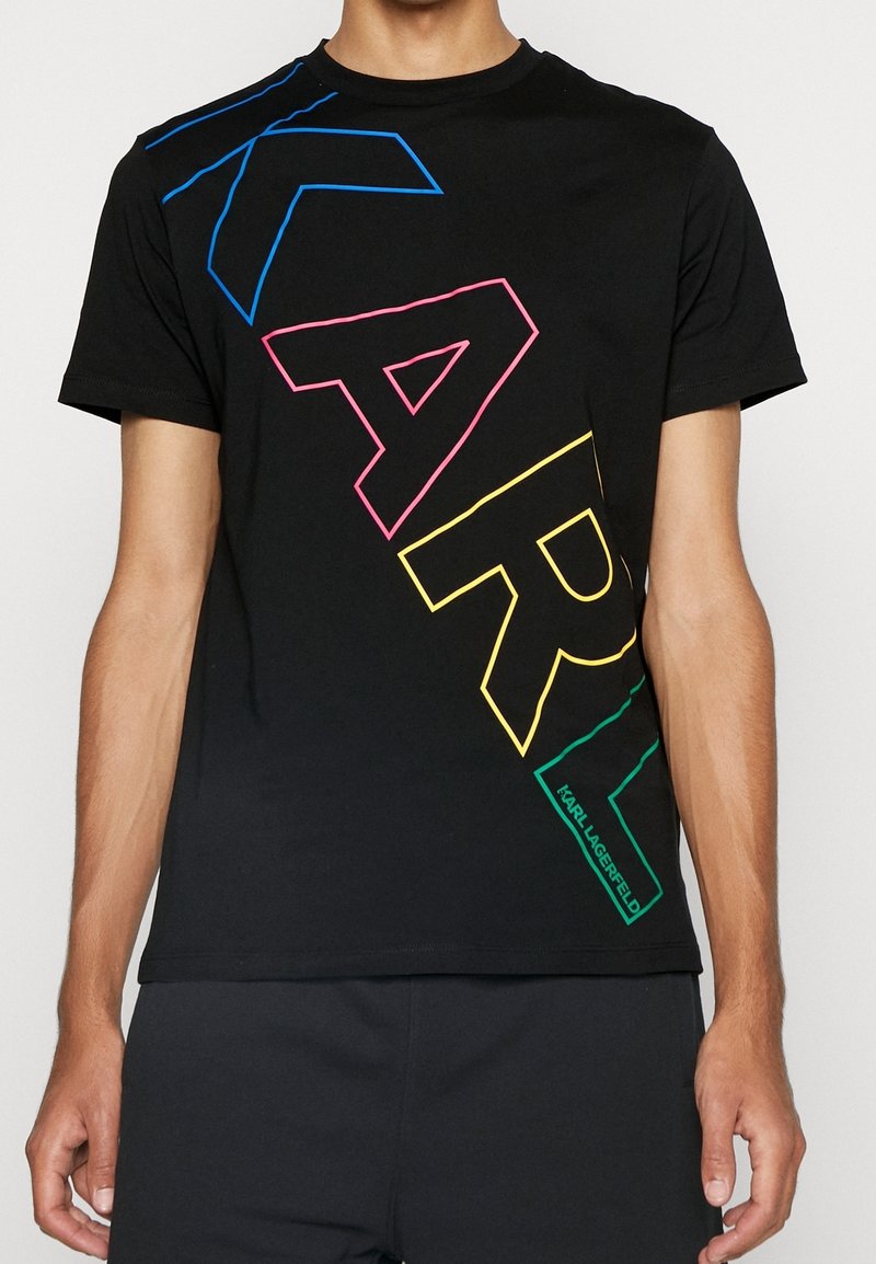 Muž v černém tričku s velkými obrysovými písmeny "KARL" v modré, růžové, žluté a zelené barvě diagonálně přes přední část.