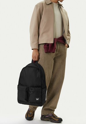 Persona con chaqueta beige y pantalones marrones sosteniendo una mochila negra de Reebok por el asa, con zapatillas multicolor y un suéter marrón atado a la cintura.