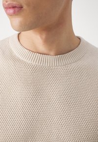 Beige stickad tröja med ett strukturerat mönster, ribbat rundhalsat nacke och mjukt tyg. Närbild som framhäver fina detaljer.