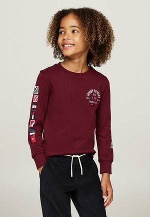 Camicia a maniche lunghe color bordeaux con logo e design di bandiere multicolori sulle maniche, abbinata a pantaloni in corduroy scuri e una coulisse bianca.