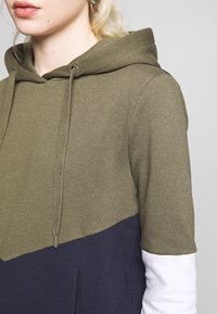 Sudadera con capucha que presenta un diseño de parte superior en verde oliva, parte inferior en azul marino y mangas blancas; confeccionada en una tela suave con un corte relajado y cordón ajustable.