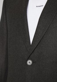 Ciemnoszary wełniany żakiet z klapami, zapinany na pojedynczy guzik, o subtelnym splocie. Biała bawełniana koszula z widocznym logo "SANDRO" poniżej.