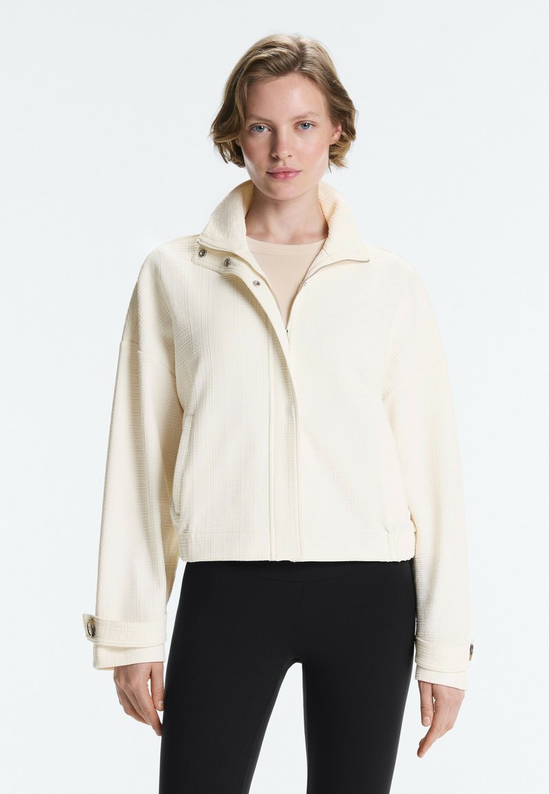 Femme portant une veste zippée crème texturée avec des boutons-pression et un pantalon noir, debout devant un fond clair uni.
