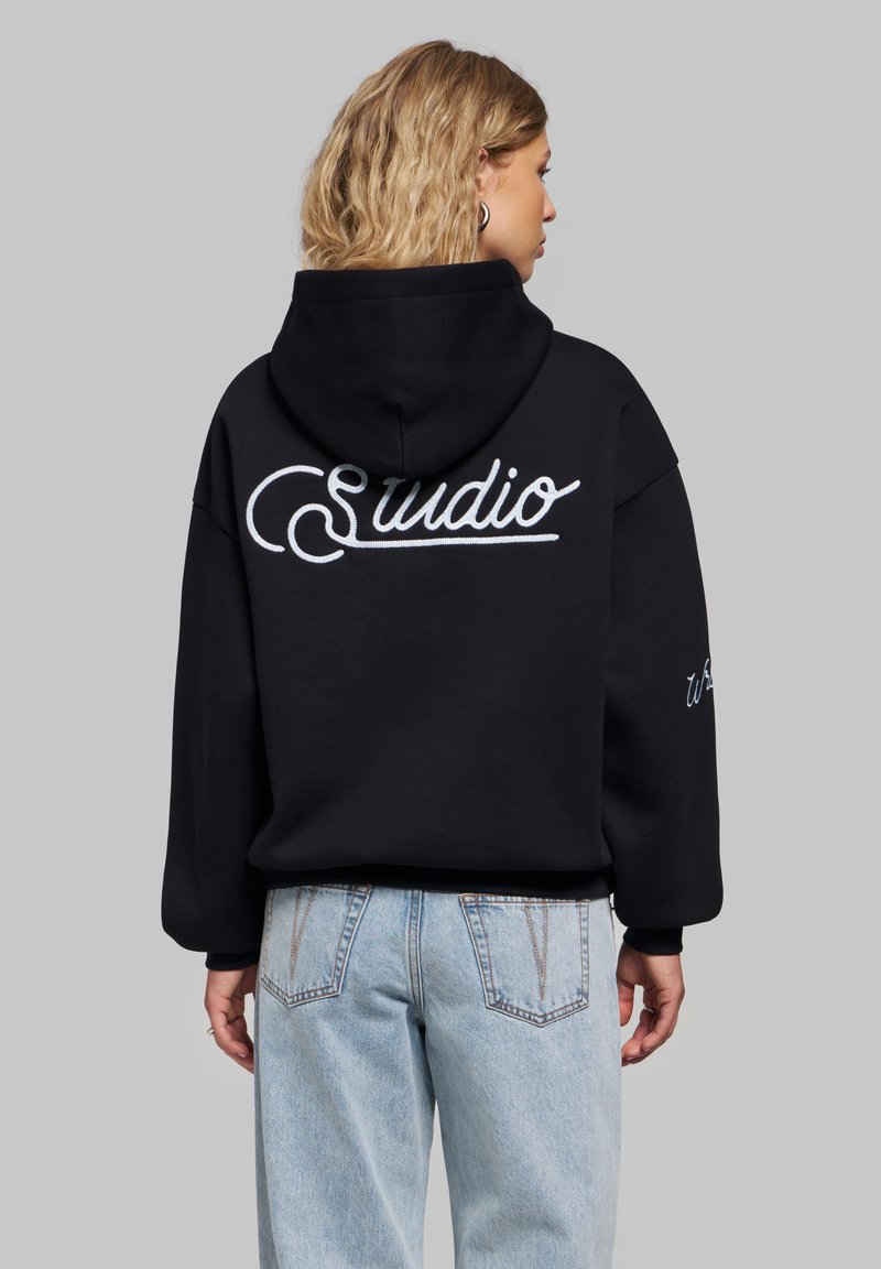 Femme aux cheveux blonds portant un sweat à capuche noir avec le texte "Sudio" dans le dos et un jean bleu clair, de dos sur fond gris.