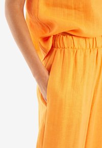 Orange linneoutfit med samlad midja, avslappnad passform och sidfickor. Slen yta med subtila färgvariationer.