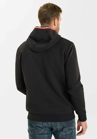 Svart hoodie med bakvy, featuring en drachordhuva, ribbade ärmslut och en trikoloraccent på kragen. Buren över blå jeans.