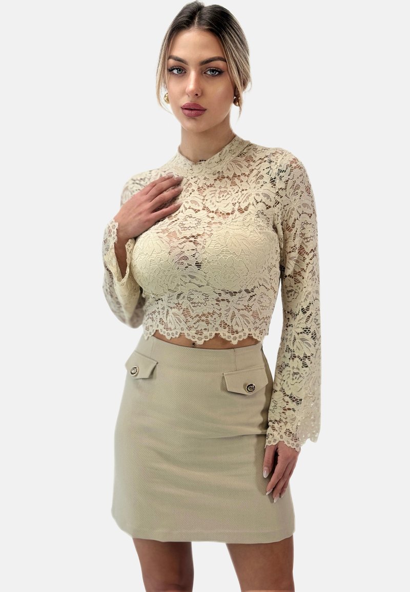 Top corto in pizzo color crema con maniche lunghe e orlo ondulato; abbinato a una gonna aderente beige con due tasche alte con bottone.