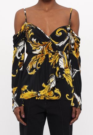 Femme portant un haut noir à manches longues, épaules dénudées, avec des motifs baroques jaune et blanc et de fines bretelles.