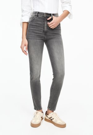 Personne portant un jean skinny gris délavé taille haute, une chemise blanche à manches retroussées et des baskets beiges avec des accents marron.