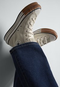 Tenisky v štýle high-top z plátna v off-white farbe, s gumovou ochrannou špičkou a hnedými gumovými akcentmi, skombinované s tmavomodrými džínsami.