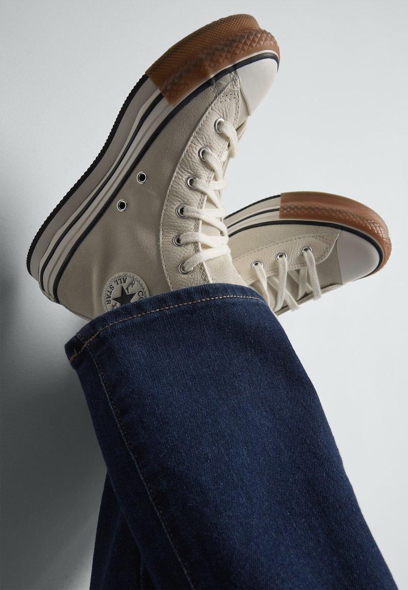 Tenisky v štýle high-top z plátna v off-white farbe, s gumovou ochrannou špičkou a hnedými gumovými akcentmi, skombinované s tmavomodrými džínsami.