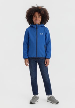 Kind mit lockigem Haar, das eine blaue Kapuzenjacke, dunkle Jeans, weiße Socken und graue Turnschuhe trägt und vor einem einfarbigen Hintergrund steht.