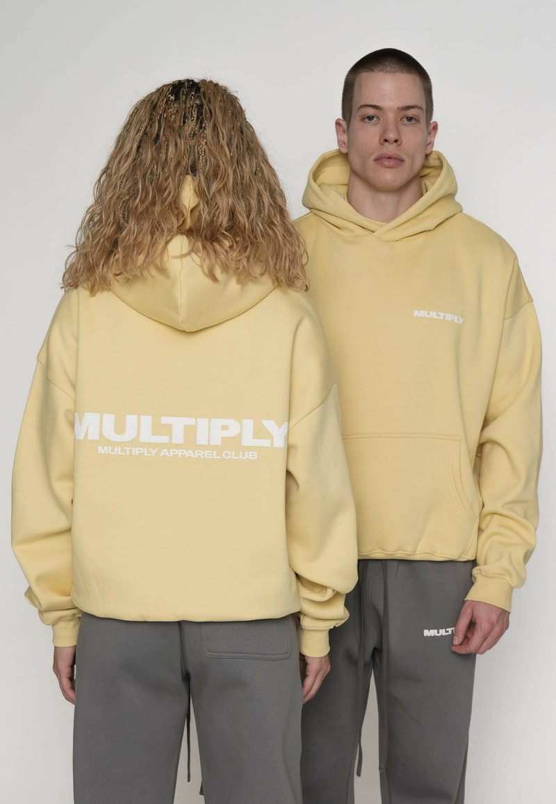 Gele hoodie met capuchon en een buidelzak aan de voorkant, met gedurfde witte tekst "MULTIPLY" op de achterkant en borst. Gecombineerd met grijze broek.