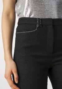 Pantalon noir texturé à coupe ajustée, avec des accents brillants le long des poches avant et de la taille.
