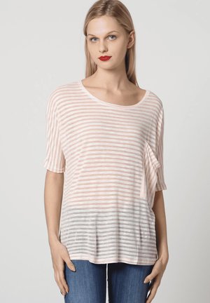 Mujer rubia con una blusa suelta de manga corta, a rayas en rosa claro y blanco, con un bolsillo en el pecho, combinada con jeans azules.