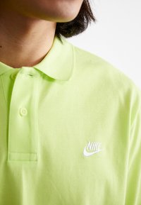 Limetkovo zelená polo košeľa Nike s golierom, s bielym vyšívaným logom na hrudi a textúrovaným povrchom látky.