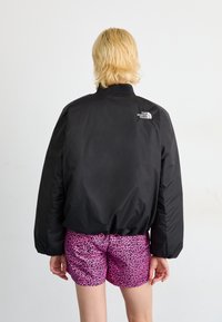 Zwarte bomberjack met hoge kraag en manchetten, voorzien van een wit logo op de linkerschouder. Gecombineerd met roze shorts met luipaardprint.