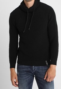 Zwarte gebreide trui met een hoge kraag, geribbeld textuur en veter aan de voorkant. Gecombineerd met blauwe jeans, met een slim fit en verwerkte details.