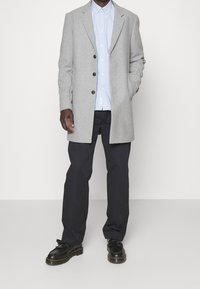 Mężczyzna w szarym płaszczu zapinanym na guziki, jasnoniebieskiej koszuli, czarnych spodniach oraz czarnych mokasynach z frędzlami, stojący na tle jednolitego jasnego tła.