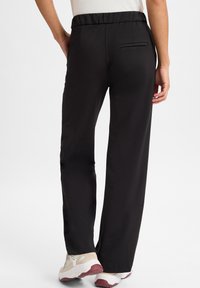 Zwarte, elastische broek met een recht model. Heeft een achterzak en een gladde, matte stof. Gecombineerd met lichte sportschoenen.