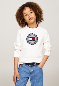 Felpa bianca con un logo circolare in blu navy e rosso. Tessuto morbido, polsini a coste, abbinata a jeans azzurri e una cintura intrecciata nera.