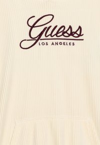 Sudadera de color crema con canalé y un bolsillo frontal, con bordado en burdeos de "Guess Los Angeles" en el pecho. Material de algodón suave.