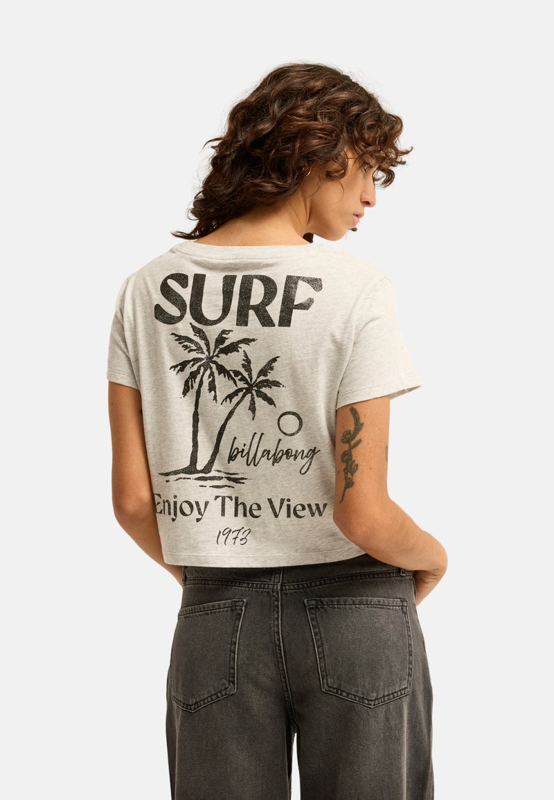 Giovane donna con capelli ricci indossa una maglietta corta grigio chiaro con palme e il testo ""SURF Enjoy The View"" sulla schiena.