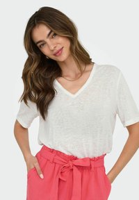 Witte t-shirt met v-hals en korte mouwen, gemaakt van lichtgewicht stof. Gepaard met hoogstaande, roze, met riem verstelbare broeken.