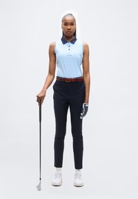 Γυναίκα όρθια με γκολφ ένδυση κρατώντας ένα γκολφ ρόπαλο, φοράει ανοιχτό μπλε αμάνικο πόλο, σκούρο μπλε παντελόνι, λευκό γάντι γκολφ και λευκά παπούτσια.