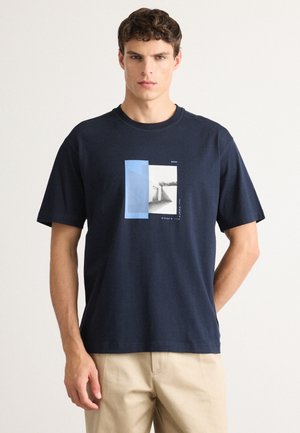 Man in een marineblauwe t-shirt met geometrische grafiek en beige broek, staand tegen een effen lichte achtergrond.