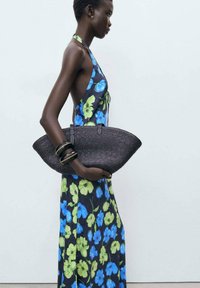 Black woven handbag with a curved shape, paired with a floral-patterned dress featuring blue and green flowers on a dark background.