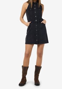 Černé džínsové šaty bez rukávů s knoflíkovým zapínáním, volánkovým pasem a bočními kapsami, doplněné hnědými kozačkami nad kolena.