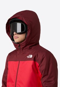 Red and maroon jacket with a hood, featuring a high collar and zipper. Model wears black ski goggles; the fabric appears smooth and weather-resistant.