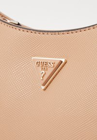 Borsa beige con texture, caratterizzata da una piastra logo dorata a forma triangolare con "GUESS USA" e punto interrogativo. Materiale simile alla pelle, design minimalista.