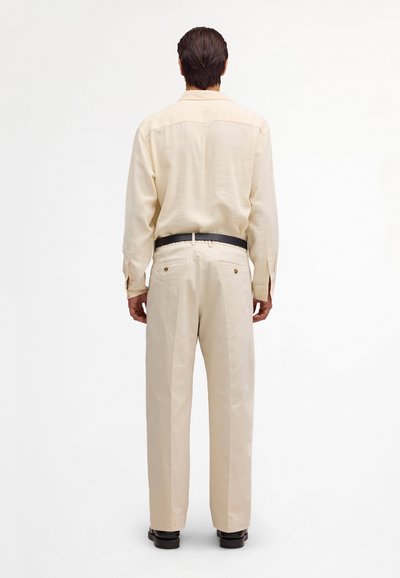 Homme debout de dos portant une chemise à manches longues crème, un pantalon crème assorti avec des poches arrière, une ceinture noire et des chaussures noires.