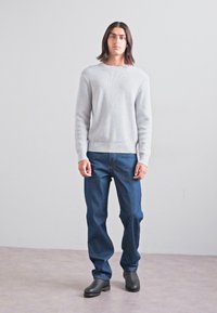 Maglione grigio a costine, jeans blu navy e stivali alla caviglia neri. La modella si trova di fronte a uno sfondo neutro, mostrando un taglio rilassato e la texture.