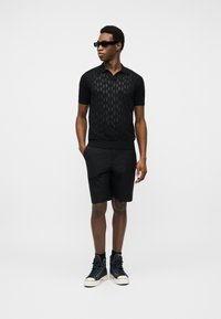 Άνδρας που φοράει μαύρο πόλο μπλουζάκι με κοντά μανίκια και σχέδιο, μαύρο σορτς, μαύρα ψηλά αθλητικά παπούτσια και σκούρα γυαλιά ηλίου, στέκεται με το ένα χέρι στην τσέπη.