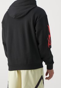 Sweat à capuche noir à texture lisse, avec un motif de flamme rouge sur la manche gauche. La coupe est décontractée, avec un ourlet qui arrive à la taille.