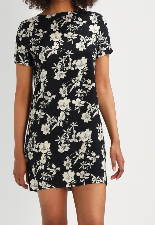 Robe courte noire à manches courtes avec imprimé floral blanc, portée par une personne à la peau de ton moyen, debout devant un fond clair uni.