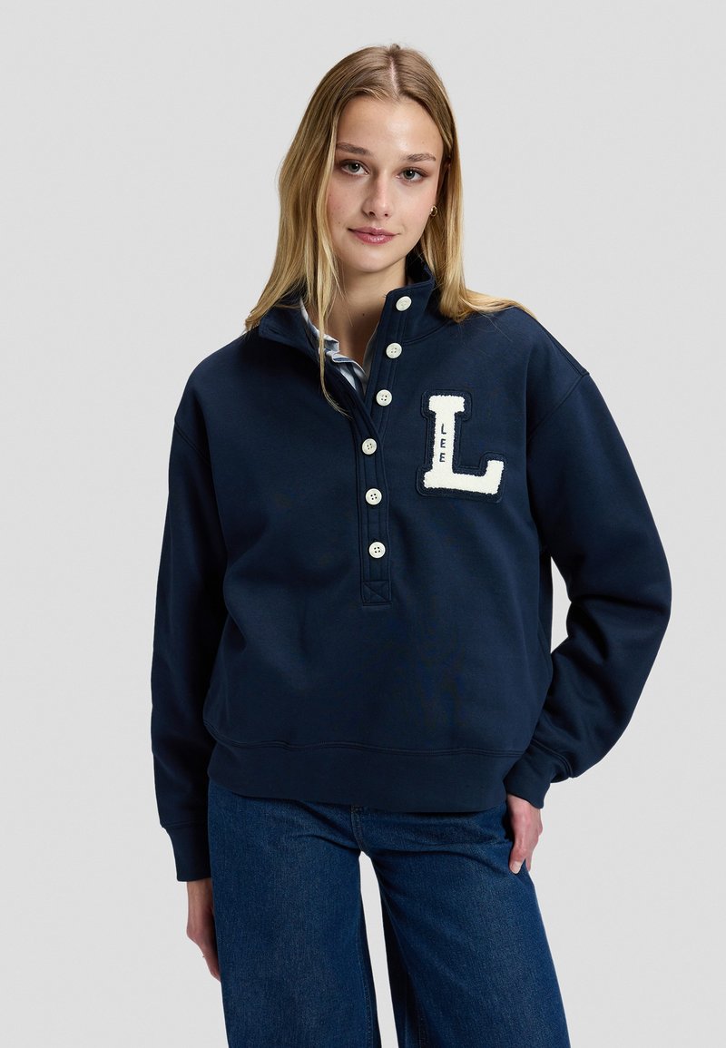 Jeune femme aux longs cheveux blonds portant un sweat bleu marine à boutons avec un patch blanc "L" et un jean bleu sur fond gris.