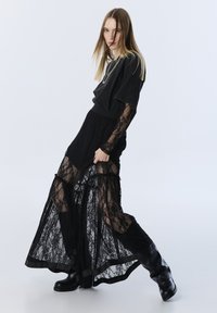 Zwarte kanten maxi-jurk met doorzichtige panelen, gelaagd over een effen zwarte top. Voorzien van lange kanten mouwen en hoge zwarte laarzen.