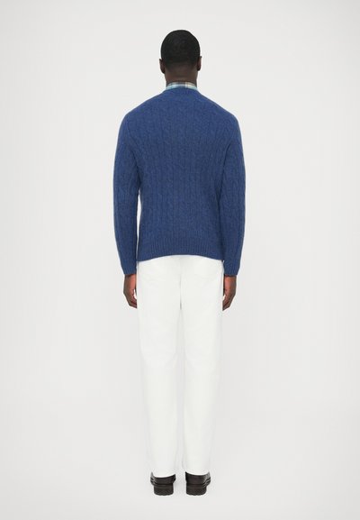 Kék kötött pulóver V-nyakkivágással, vastag textúrával és bordázott mandzsettával, fehér nadrággal és barna cipővel párosítva, hátulnézetből.