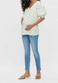Blusa floreale gialla chiara con maniche a puff, abbinata a jeans blu aderenti e sandali color tan. La blusa presenta un collo a V e dettagli sottili.