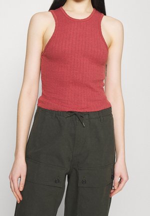 Femme portant un débardeur rouge côtelé sans manches et un pantalon vert foncé avec des poches avant boutonnées, debout devant un fond clair.