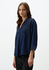 Blusa blu navy con collare, caratterizzata da ricami floreali e dettagli di perline su una spalla. Maniche lunghe con design a polsino arrotolato.